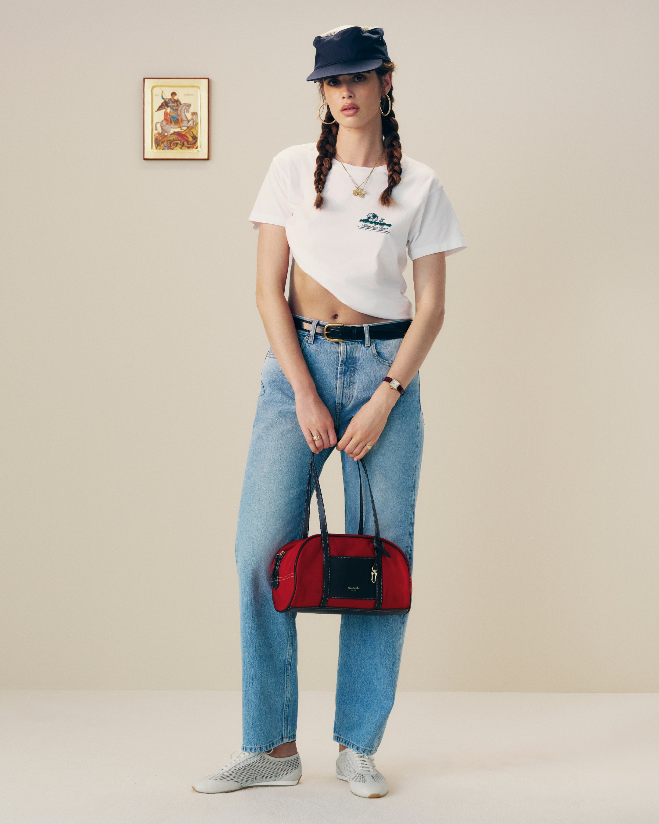 Woman in a white crop tee, jeans, and cap holds the Mini Duffle Shoulder Bag in red, standing by wall art.