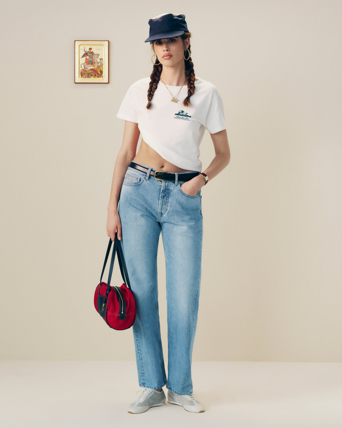 A woman in a Cropped Aloe-Blend Unisphere Tee, blue jeans, cap, and sneakers holds a red bag against a plain background.