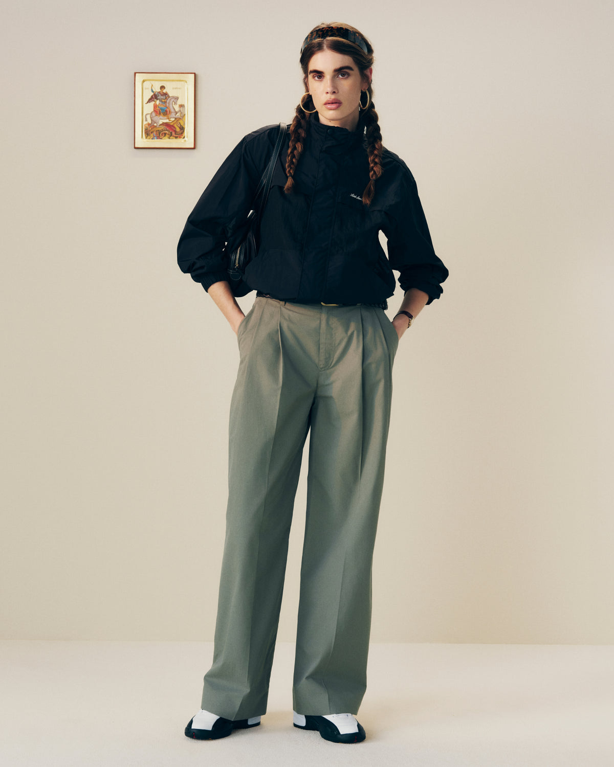 Person with braided hair wears black blouse, Lightweighted Pleated Cotton Trouser, and black-and-white shoes in a minimalist room.