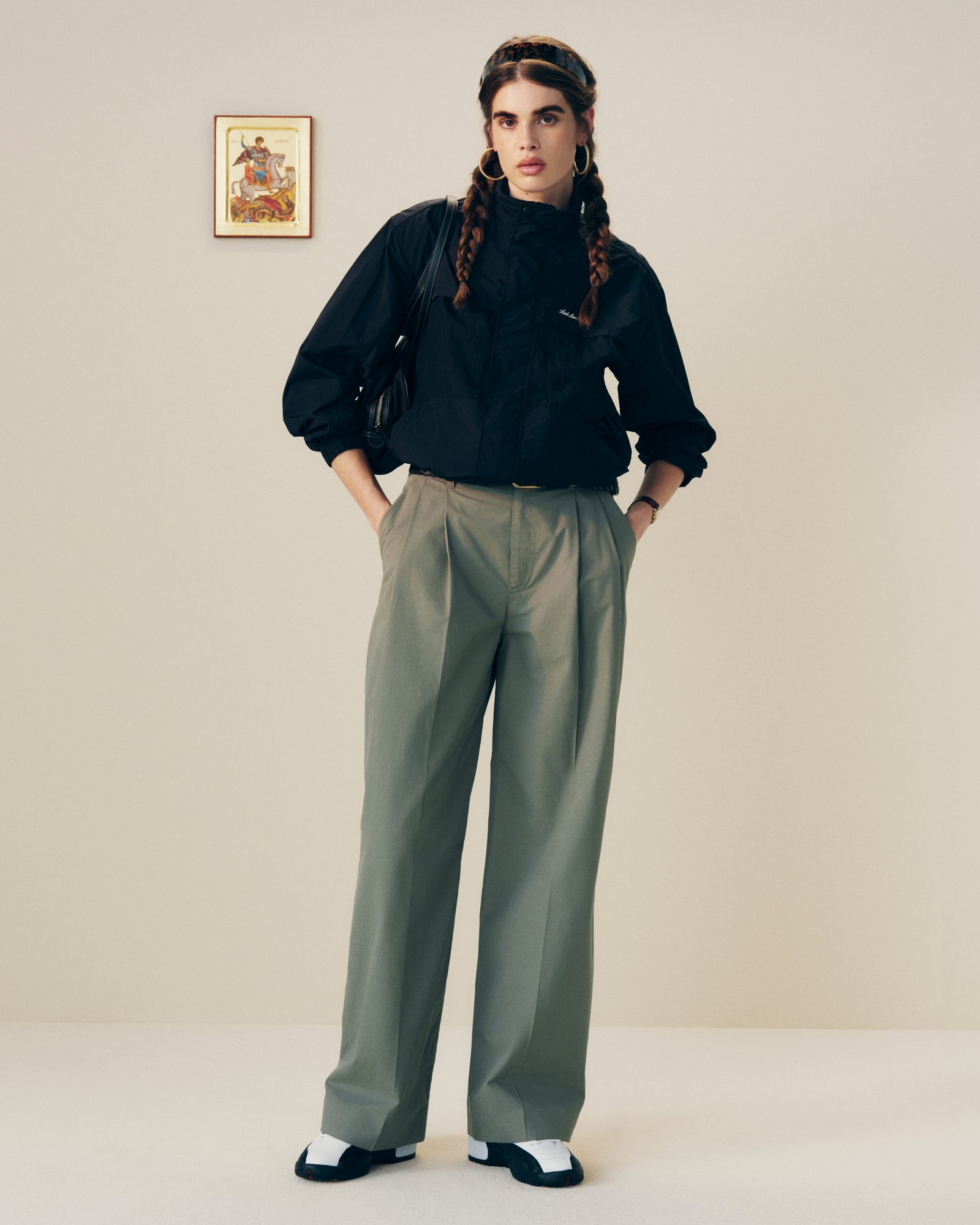 Person with braided hair wears black blouse, Lightweighted Pleated Cotton Trouser, and black-and-white shoes in a minimalist room.
