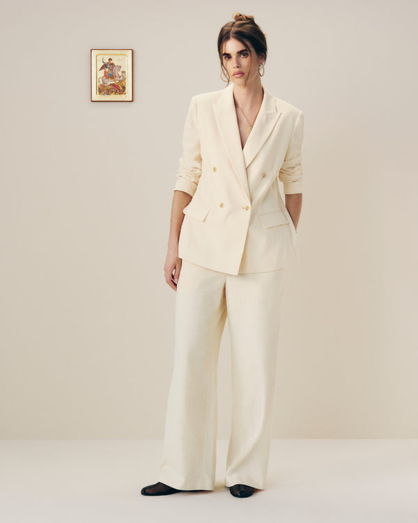 A woman models Drawstring Cotton Blend Suit Trousers in cream against a plain wall with framed art behind her.