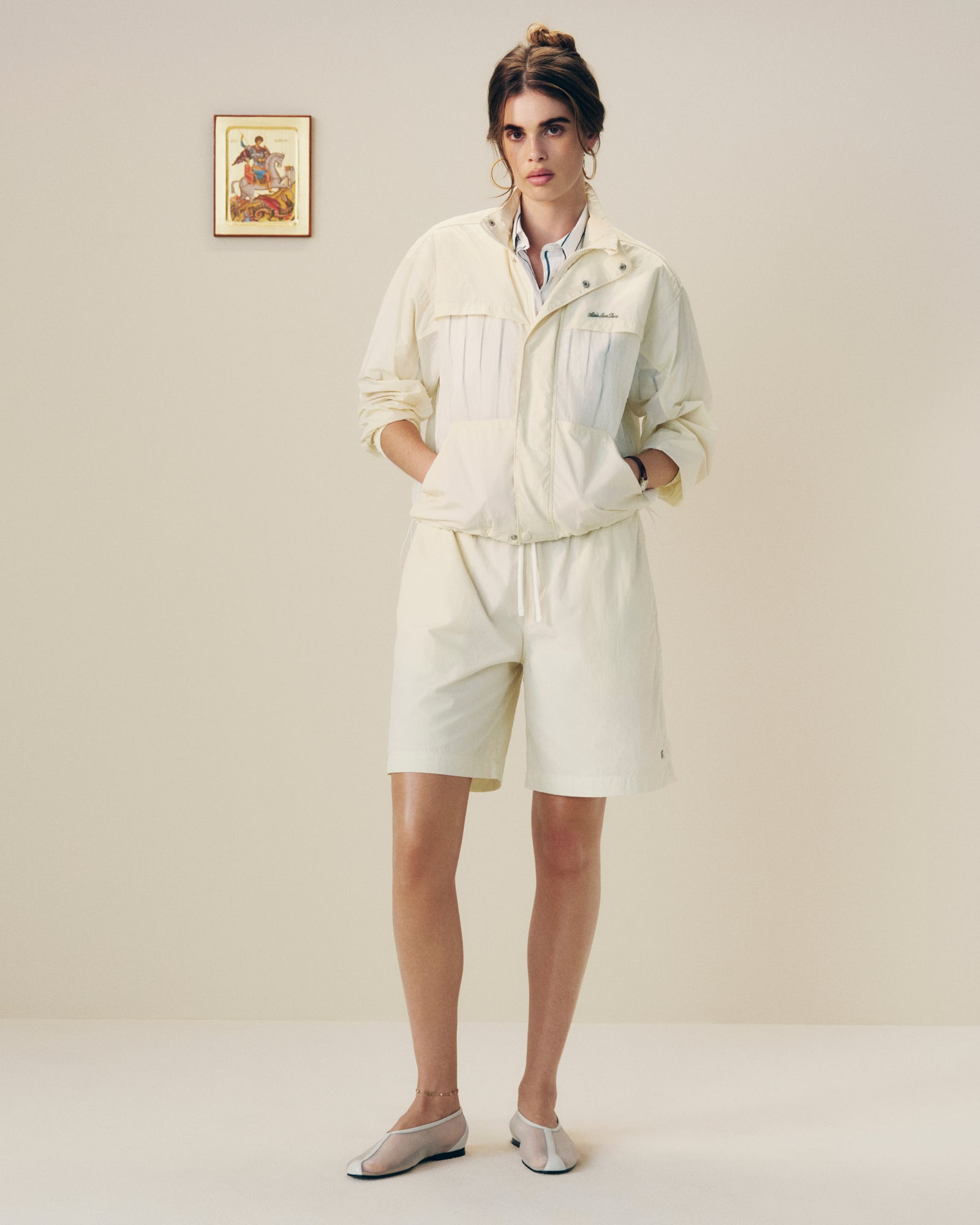 A woman in a Nylon Track Jacket and shorts stands indoors with hands in pockets near a framed wall picture.