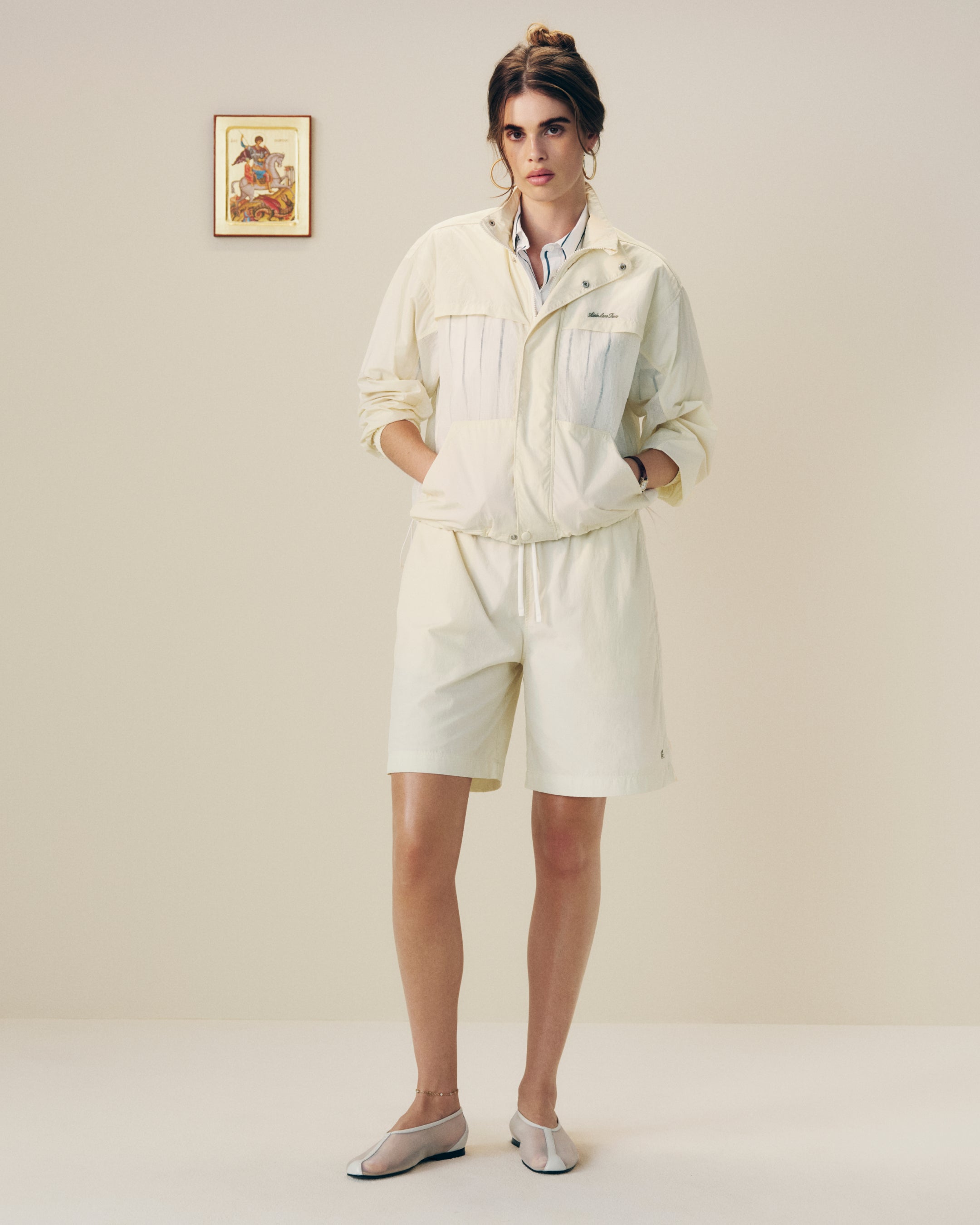 A woman in a Nylon Track Jacket and shorts stands indoors with hands in pockets near a framed wall picture.