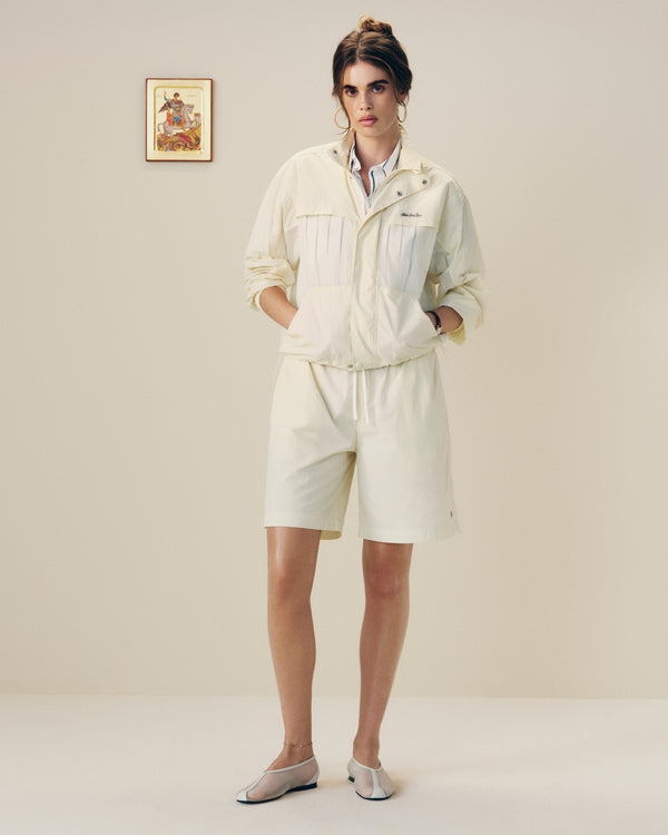 A woman in a Nylon Track Jacket and shorts stands indoors with hands in pockets near a framed wall picture.
