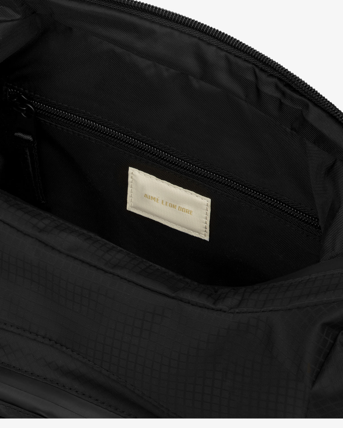 Close-up of the Nylon Messenger Bag's black interior with a beige Aimé Leon Dore label above a zippered pocket.