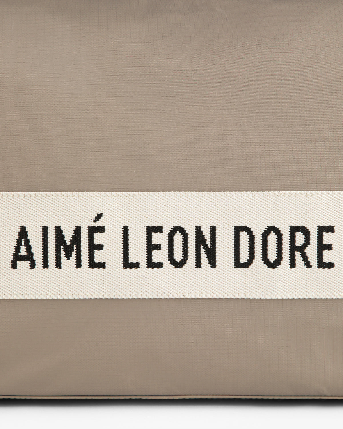 A beige nylon duffle bag with a white band featuring AIMÉ LEON DORE in black capital letters.