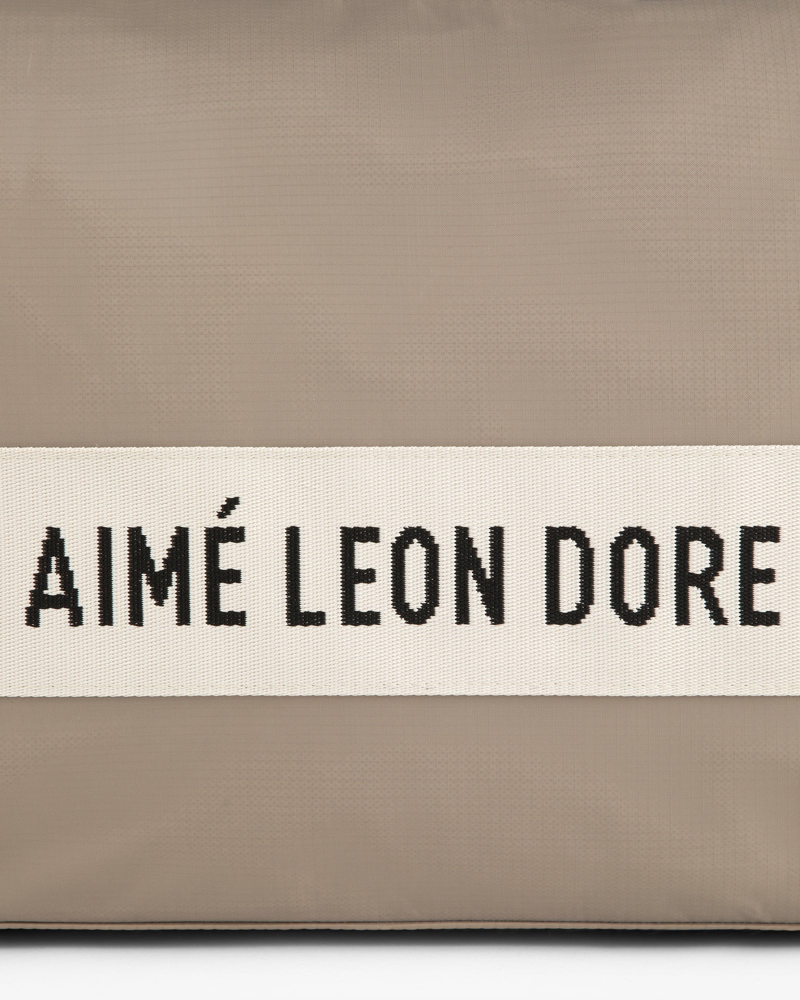A beige nylon duffle bag with a white band featuring AIMÉ LEON DORE in black capital letters.