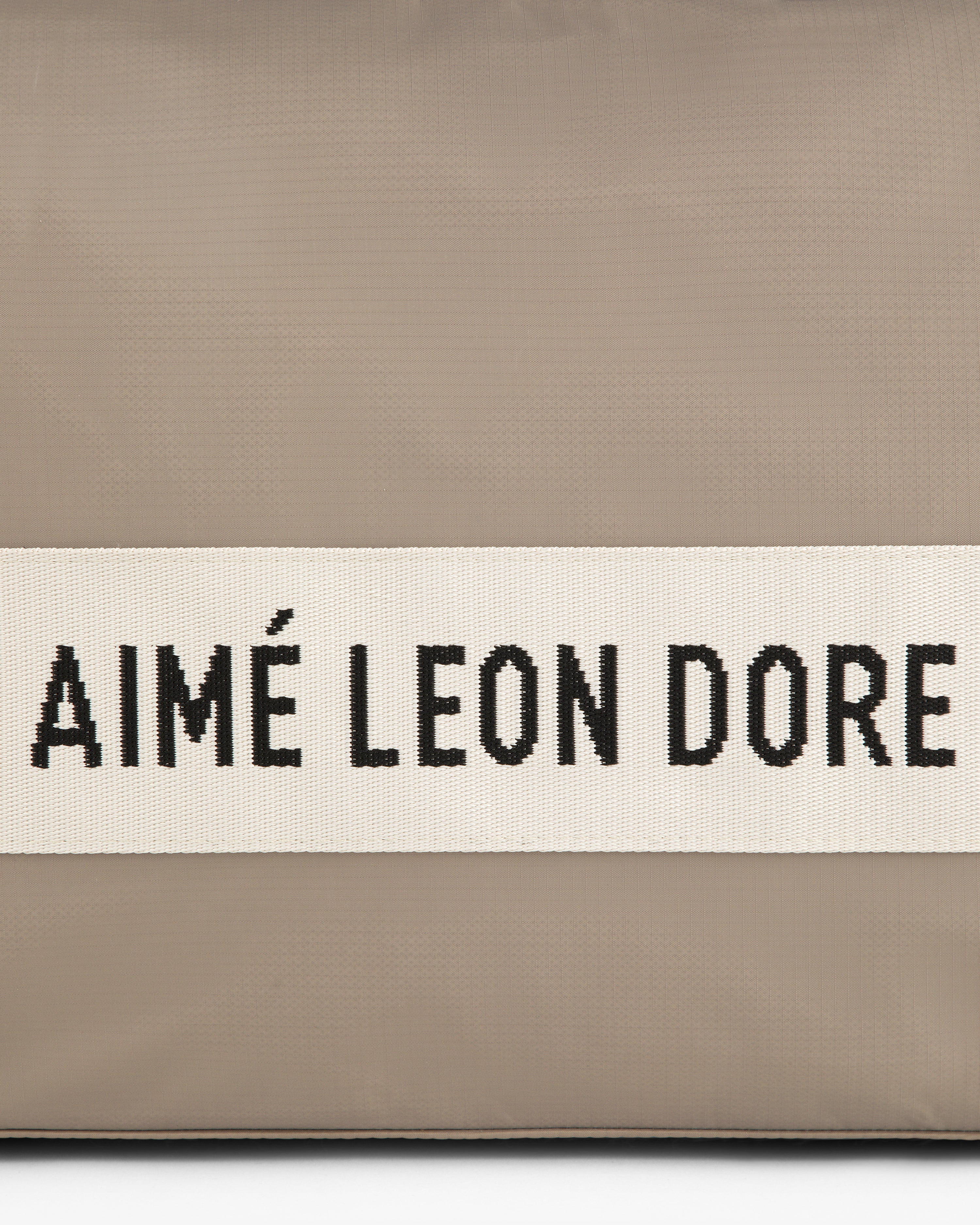 A beige nylon duffle bag with a white band featuring AIMÉ LEON DORE in black capital letters.