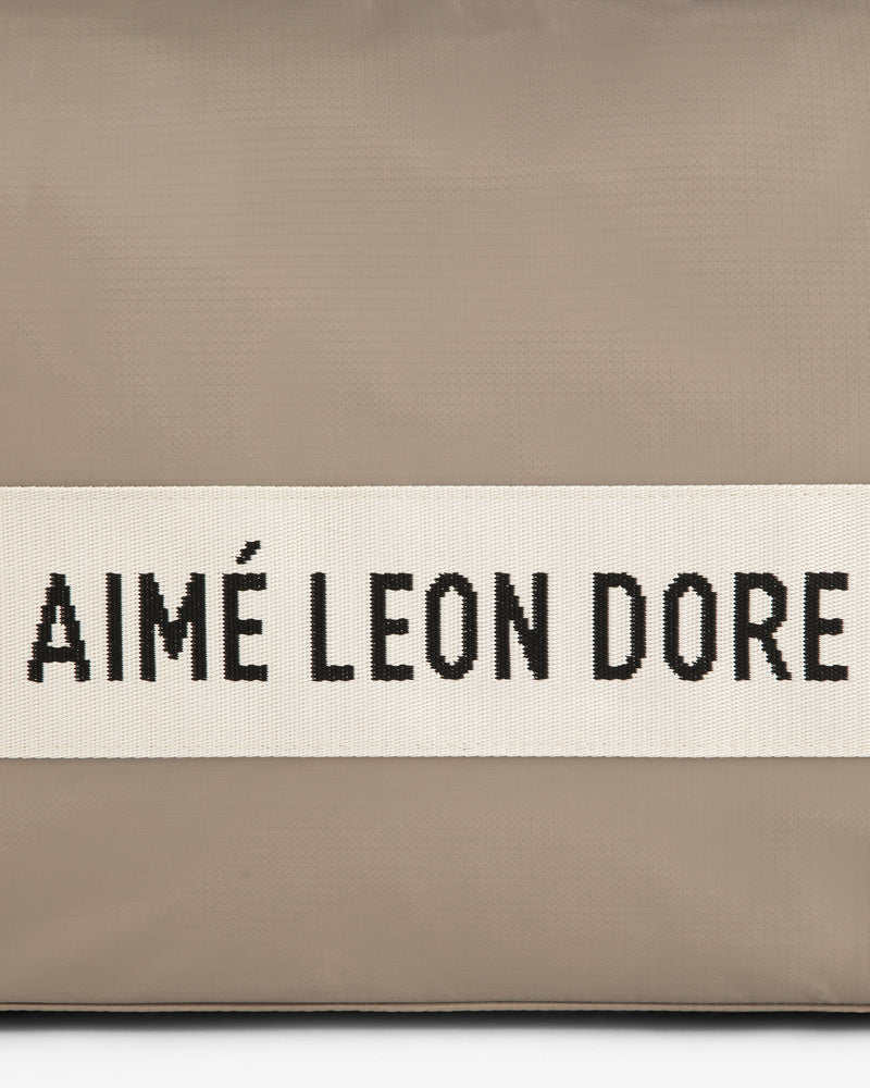 A beige nylon duffle bag with a white band featuring AIMÉ LEON DORE in black capital letters.