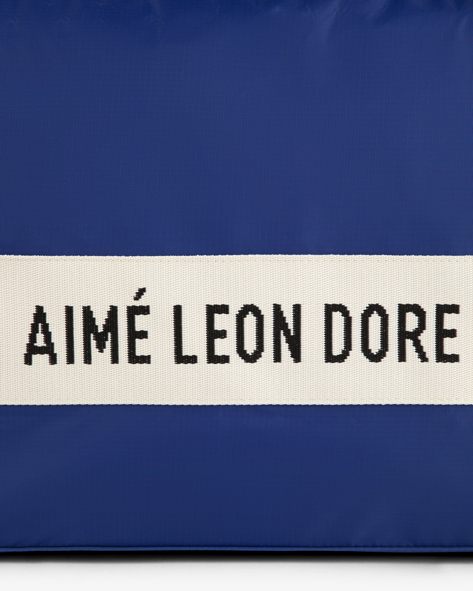A blue nylon duffle bag with a white strap and black AIMÉ LEON DORE text.