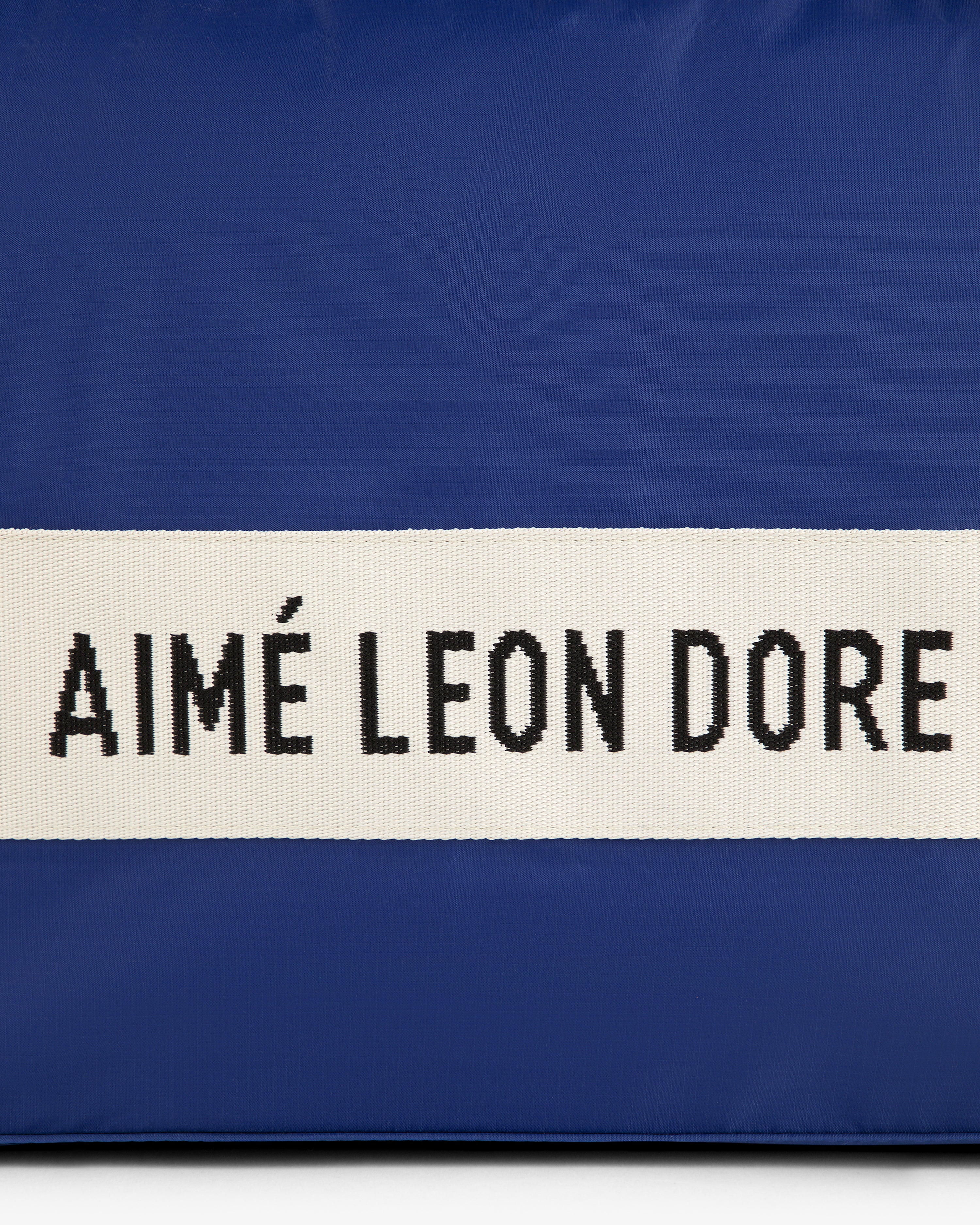 A blue nylon duffle bag with a white strap and black AIMÉ LEON DORE text.