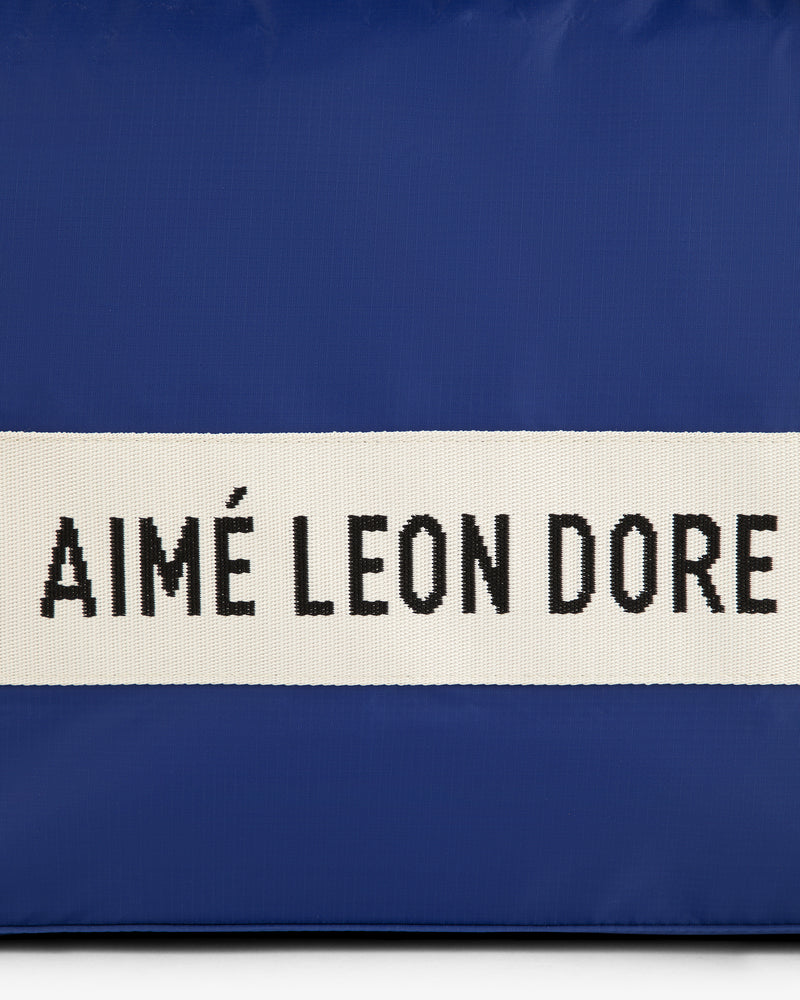 A blue nylon duffle bag with a white strap and black AIMÉ LEON DORE text.