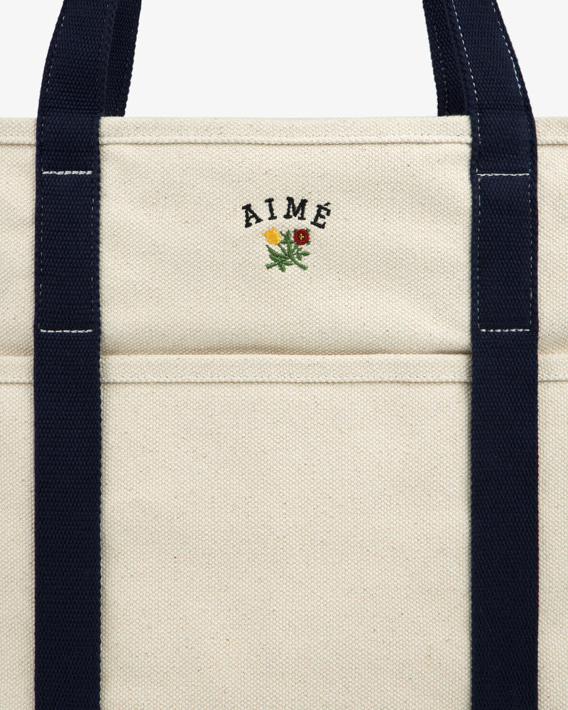 Close-up of the Large Crest Contrast Tote Bag with navy handles and AIMÉ embroidered above a small yellow-red flower.