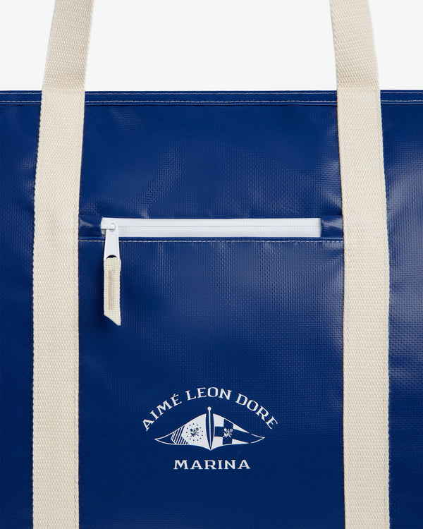 Carryall Marina Tote: Blue bag with white handles, front zip pocket, Aimé Leon Dore Marina text, and flag graphic.