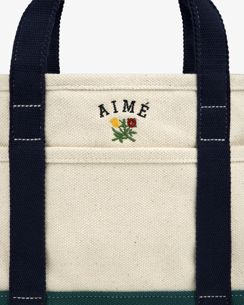 Mini Crest Contrast Tote Bag: cream canvas with navy handles, AIMÉ text, and yellow-red flower above the pocket.