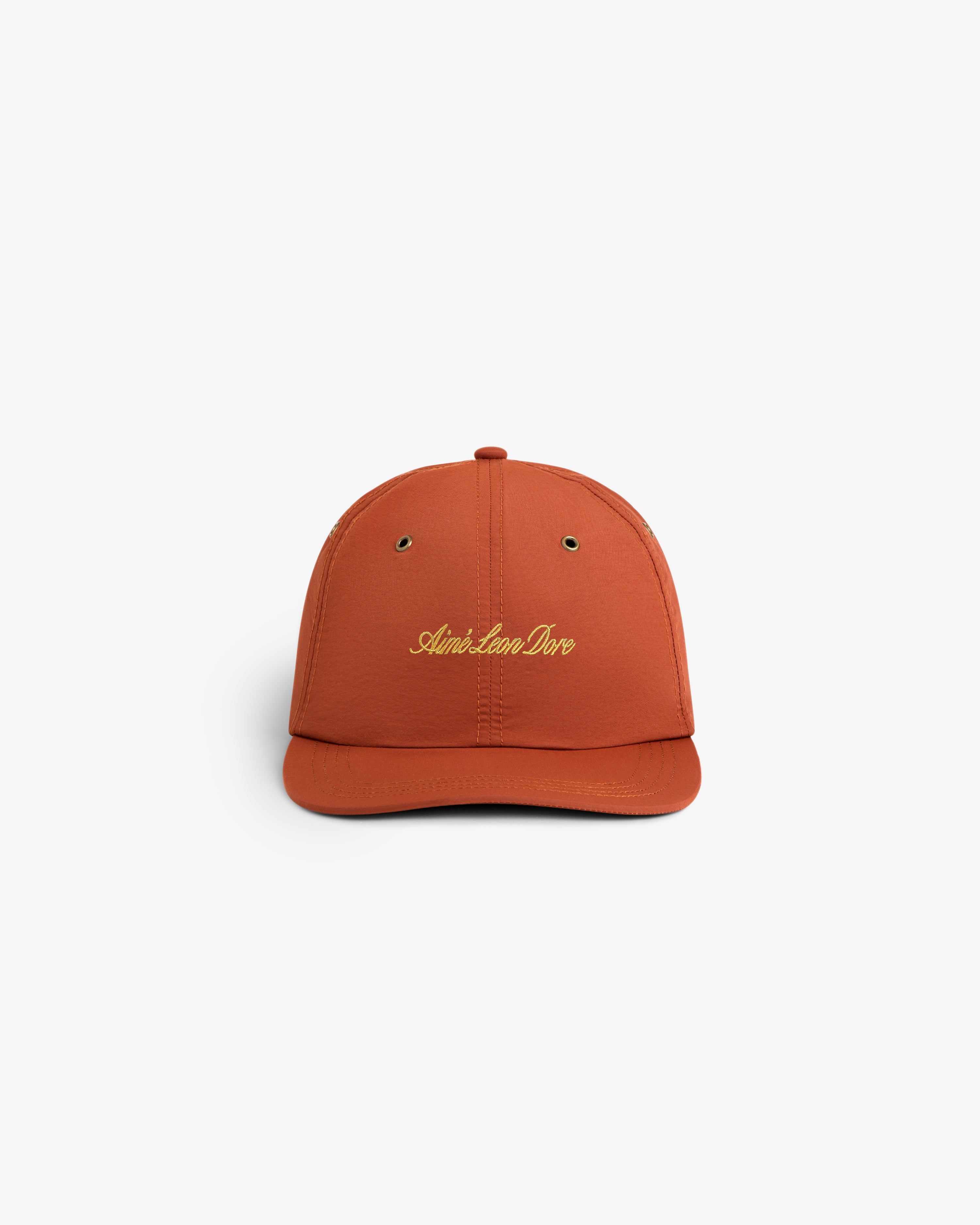 Rust-colored Script Sport Hat with Aimé Leon Dore in yellow embroidery, shown against a white background.