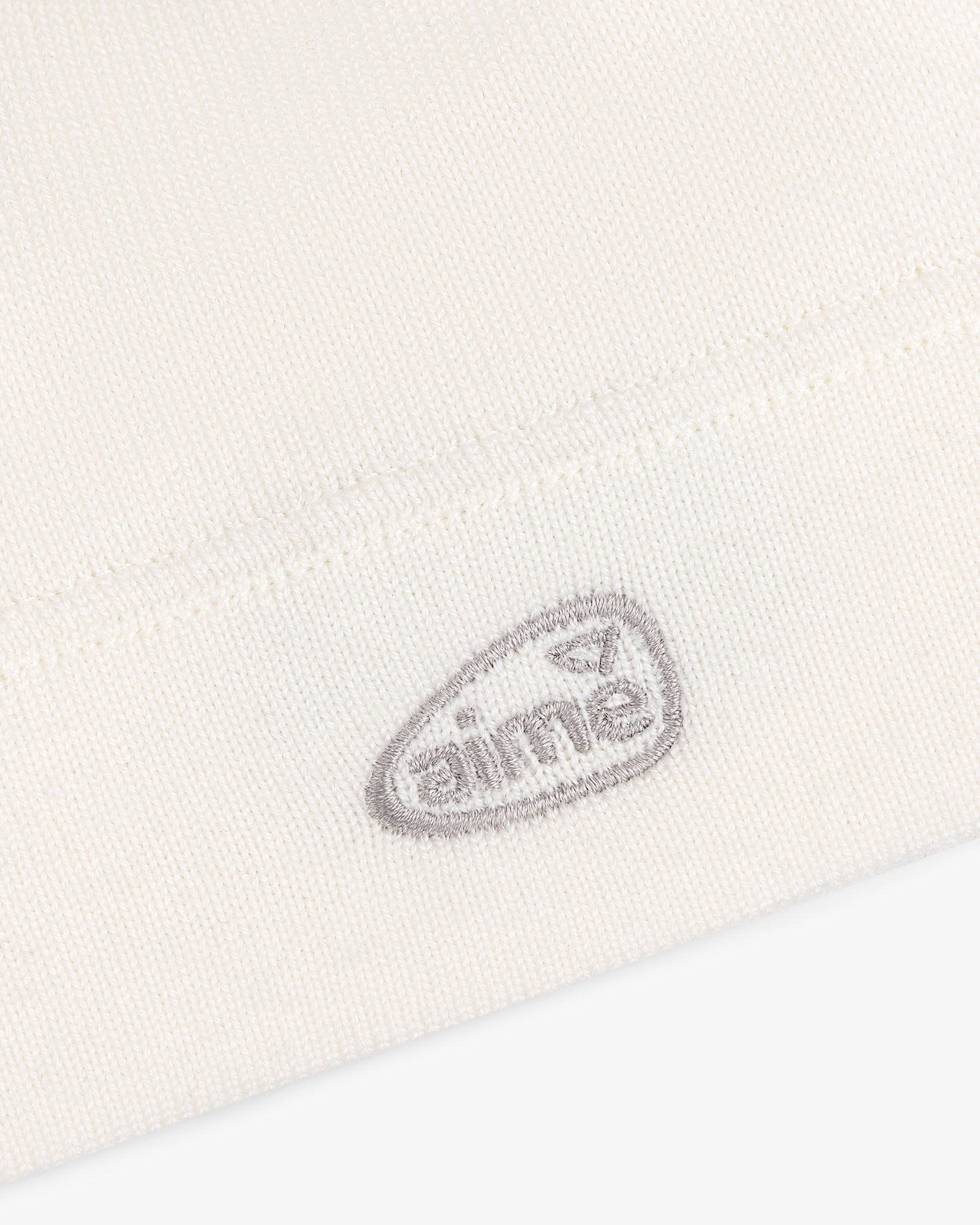 Close-up of the ALD Golf Skull Cap's white knit fabric with a small gray aimé logo embroidered near the hem.