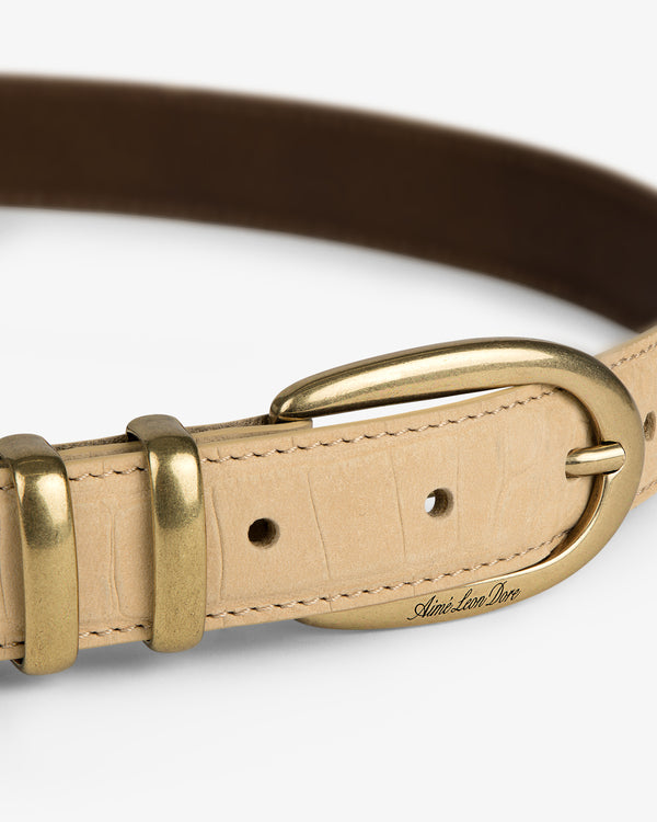 The Croc-Embossed Nubuck Belt is beige with a gold buckle, two loops, and engraved text near the buckle.