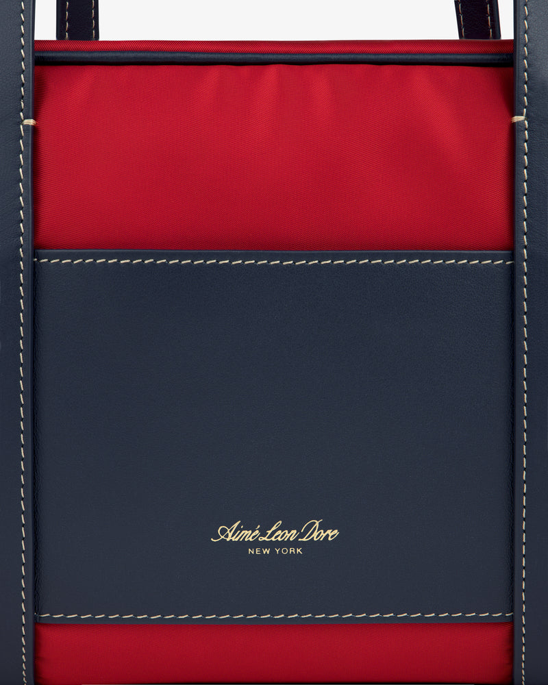Close-up of a red and navy Mini Duffle Shoulder Bag with white stitching and gold Aimé Leon Dore New York text.