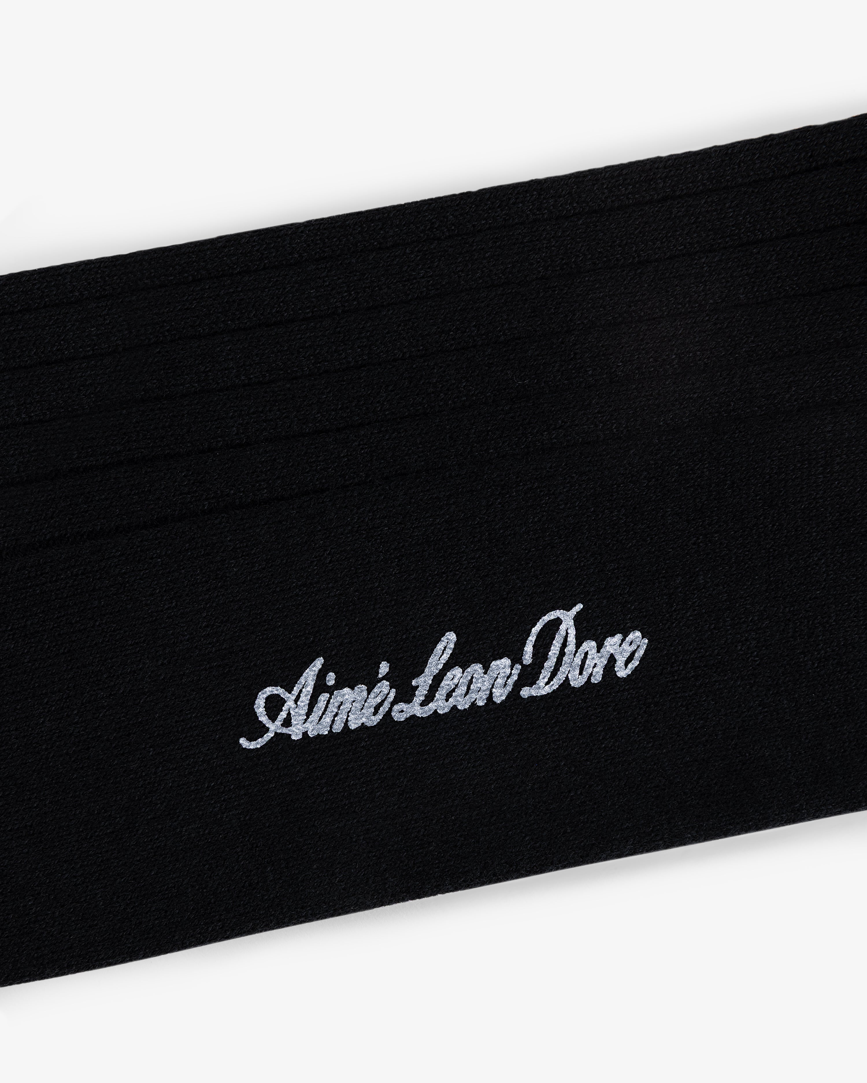 Close-up of black Dress Sock with Aimé Leon Dore embroidered in white cursive text.