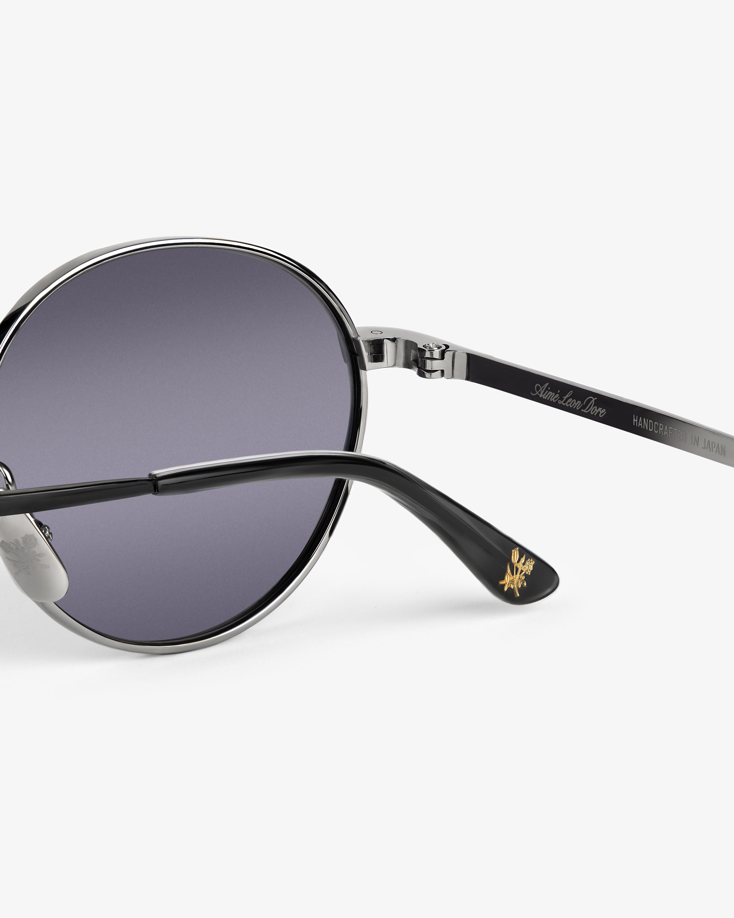 Close-up of Giórgios Sunglasses with a silver frame, purple lenses, and gold logo on the black arm tip.