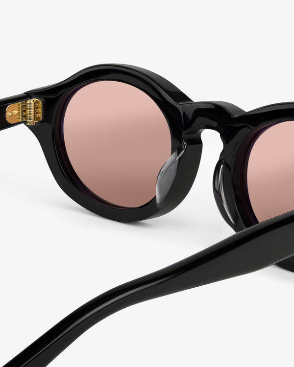 Close-up of Onásis Sunglasses—black, round frame with pink-tinted lenses against a white background.