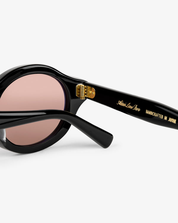 Close-up of Onásis Sunglasses: black frames, pink lenses, and "Handcrafted in Japan" gold text on the arm.