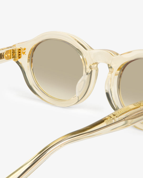 Close-up of Onásis Sunglasses: round, clear frames with gold accents and tinted lenses on a white background.