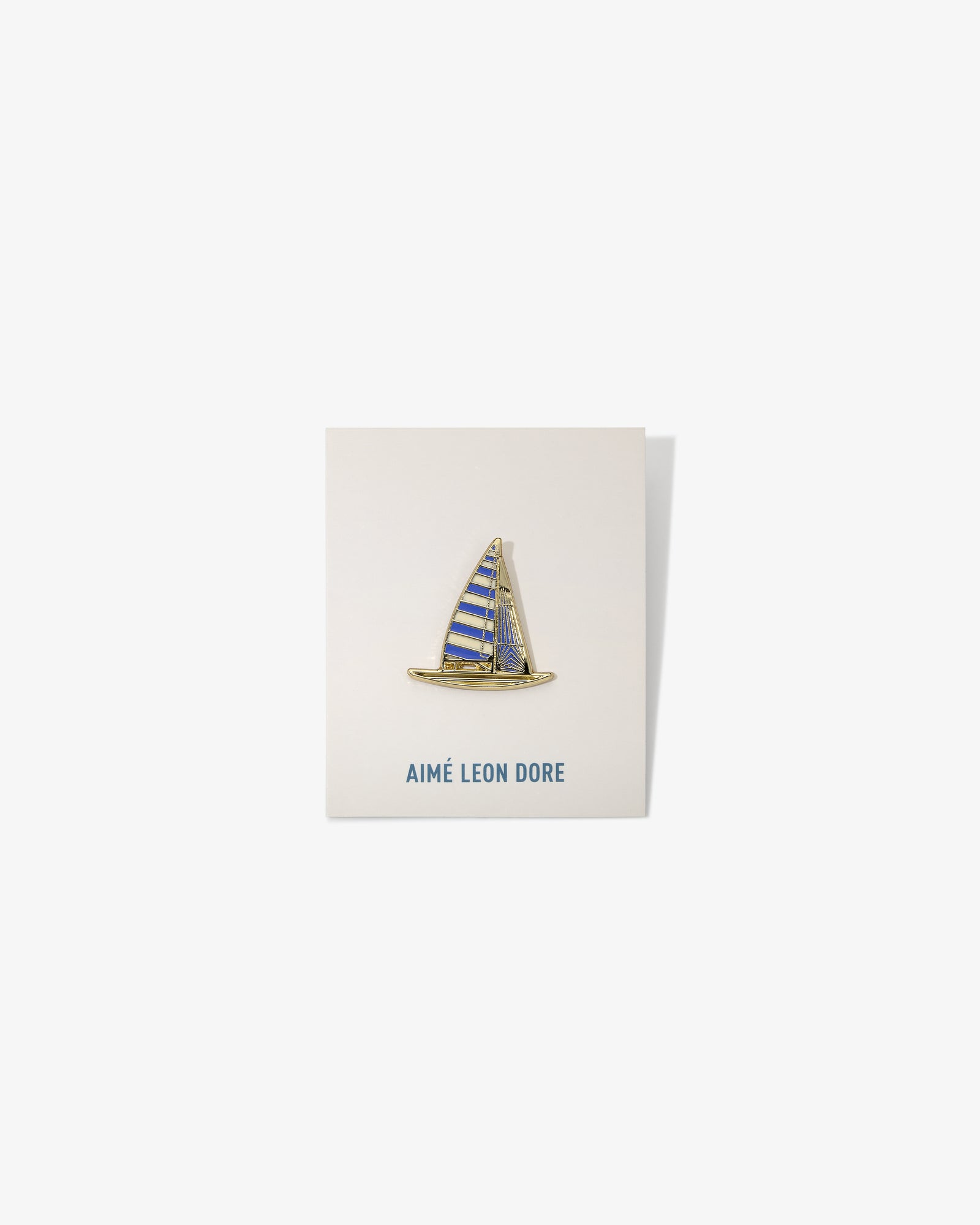 A gold and blue Graphic Pin shaped like a sailboat on a white AIMÉ LEON DORE card.