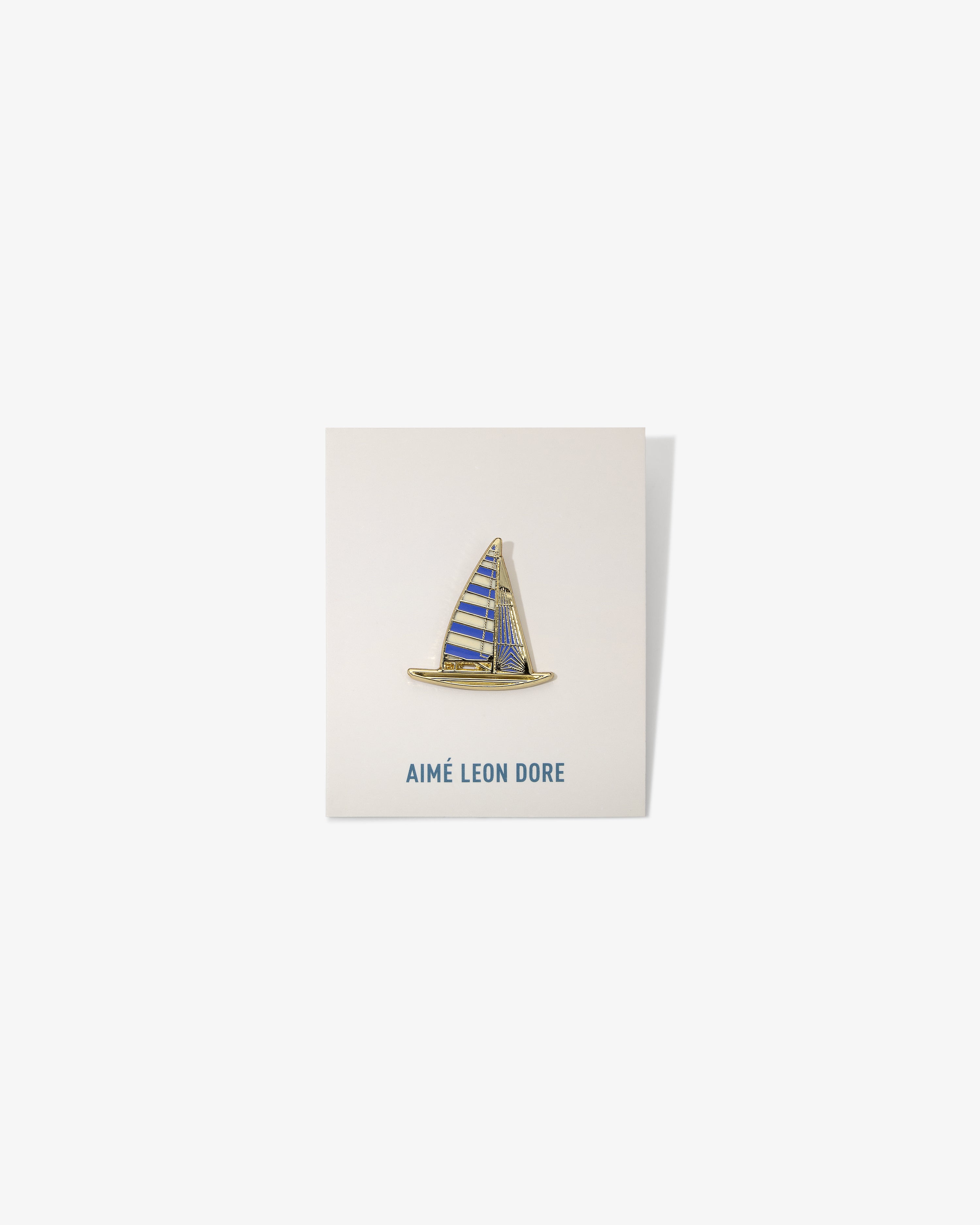 A gold and blue Graphic Pin shaped like a sailboat on a white AIMÉ LEON DORE card.