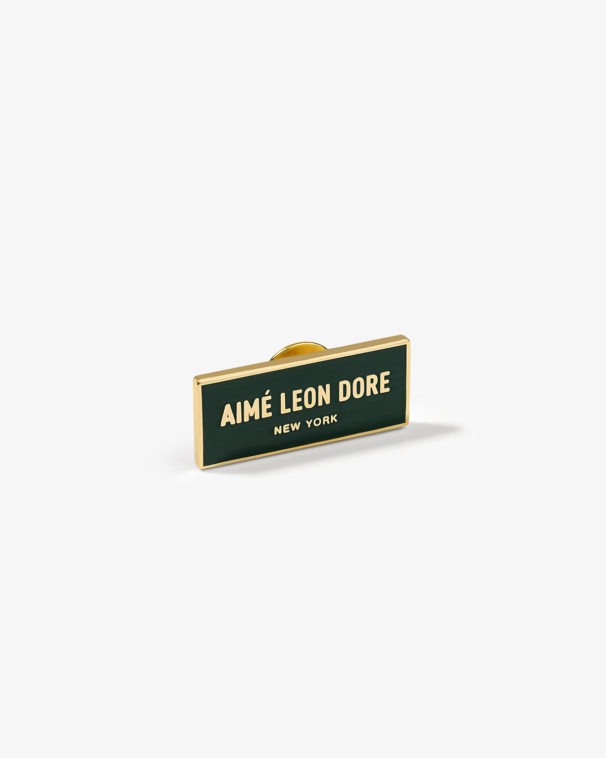 The Graphic Pin is a rectangular enamel pin with AIMÉ LEON DORE NEW YORK in gold on a dark green background.