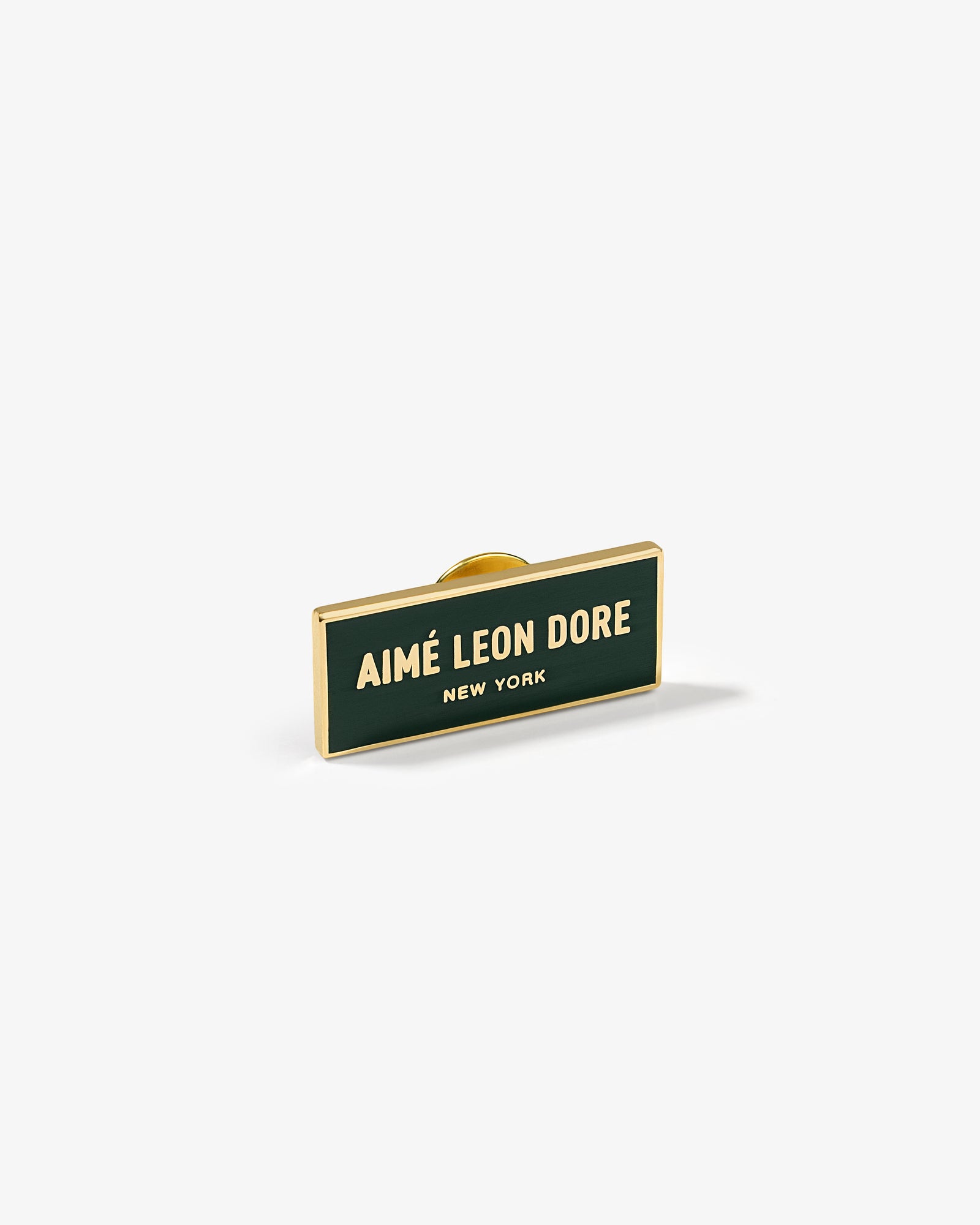 The Graphic Pin is a rectangular enamel pin with AIMÉ LEON DORE NEW YORK in gold on a dark green background.