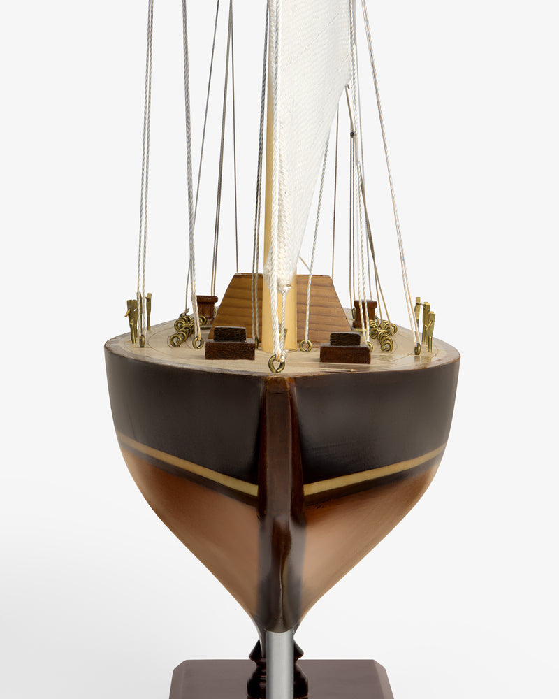 Walnut Model Sailboat viewed from the front, featuring a brown hull, deck details, and a white sail with rigging.