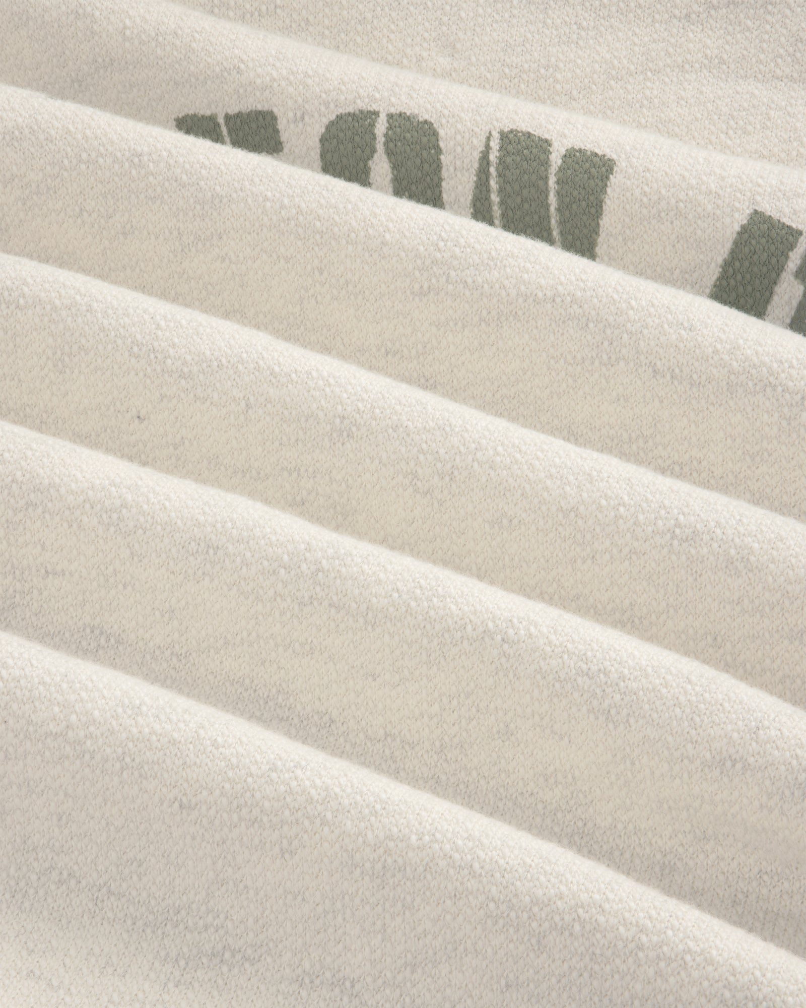 Close-up of Dock Hoodie’s light fabric with faint green text, showing soft texture and horizontal folds.