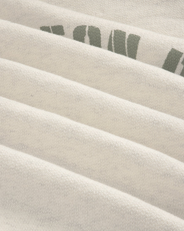 Close-up of Dock Hoodie’s light fabric with faint green text, showing soft texture and horizontal folds.