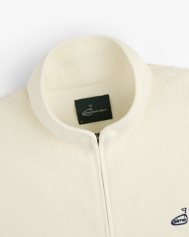 Close-up of the cream ALD Golf Piqué Full Zip Sweater with textured fabric, stand collar, and inner Aimé Leon Dore label.