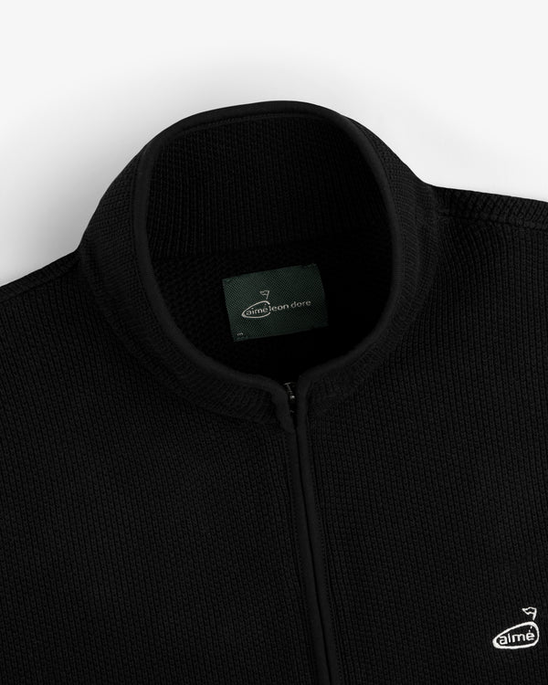 Close-up of an ALD Golf Piqué Full Zip Sweater in black with label and small embroidered chest logo.