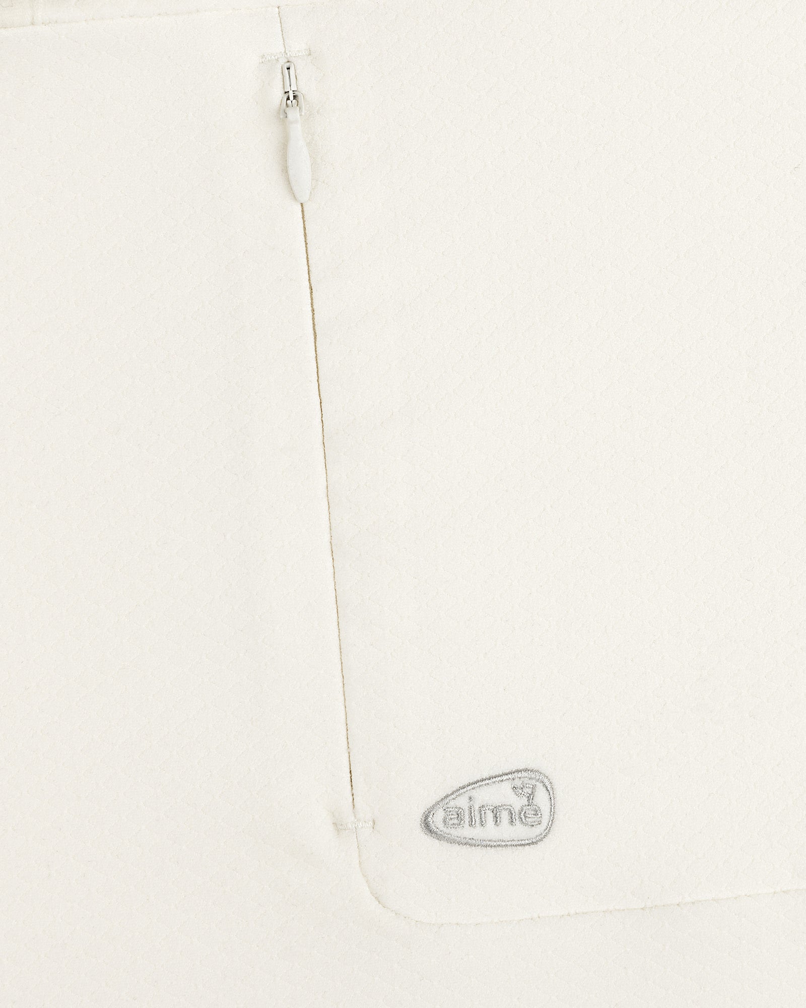 Close-up of the ALD Golf Quarter Zip Pullover’s white textured fabric with a zipper and gray “aimé” logo near the seam.