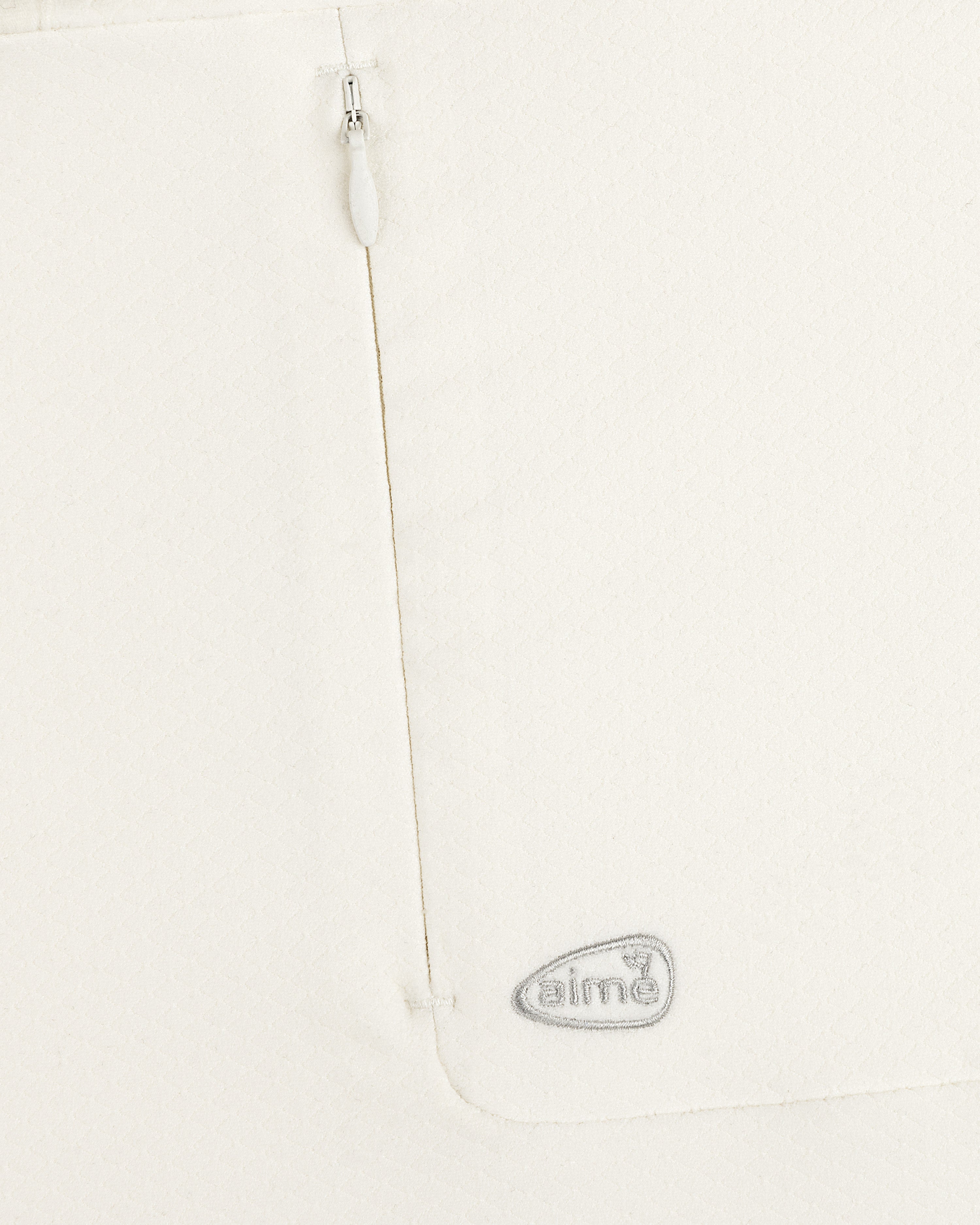 Close-up of the ALD Golf Quarter Zip Pullover’s white textured fabric with a zipper and gray “aimé” logo near the seam.