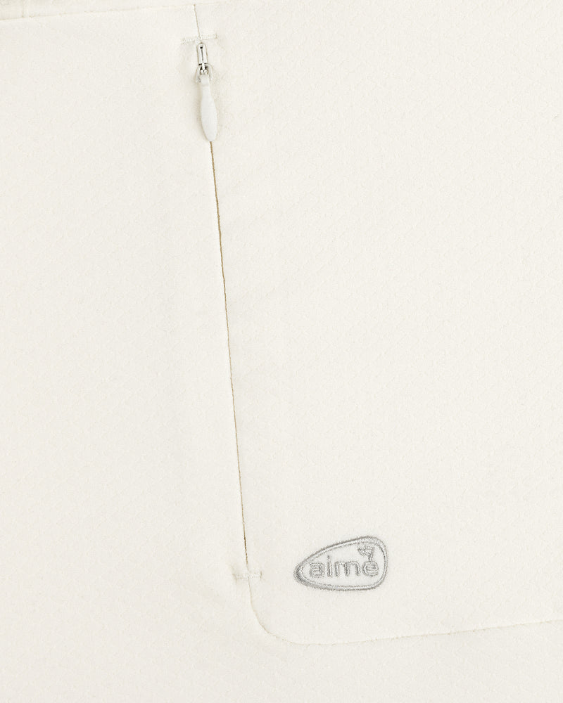 Close-up of the ALD Golf Quarter Zip Pullover’s white textured fabric with a zipper and gray “aimé” logo near the seam.