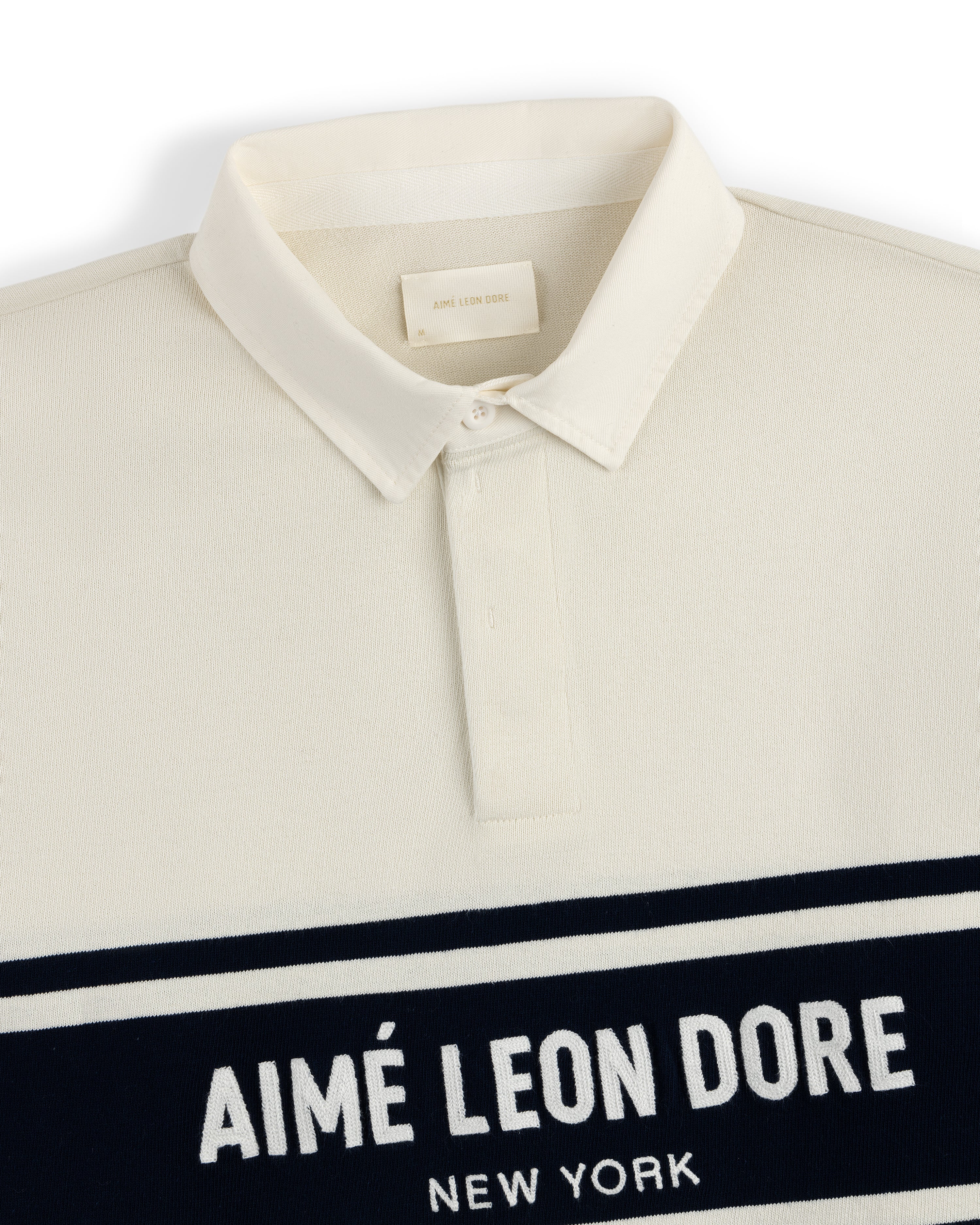 Chest Stripe Logo Rugby with a cream base, collar, and AIMÉ LEON DORE NEW YORK text on a navy chest stripe.