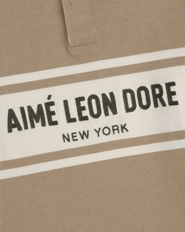 Close-up of a beige Chest Stripe Logo Rugby with AIMÉ LEON DORE NEW YORK in bold black on a white chest stripe.