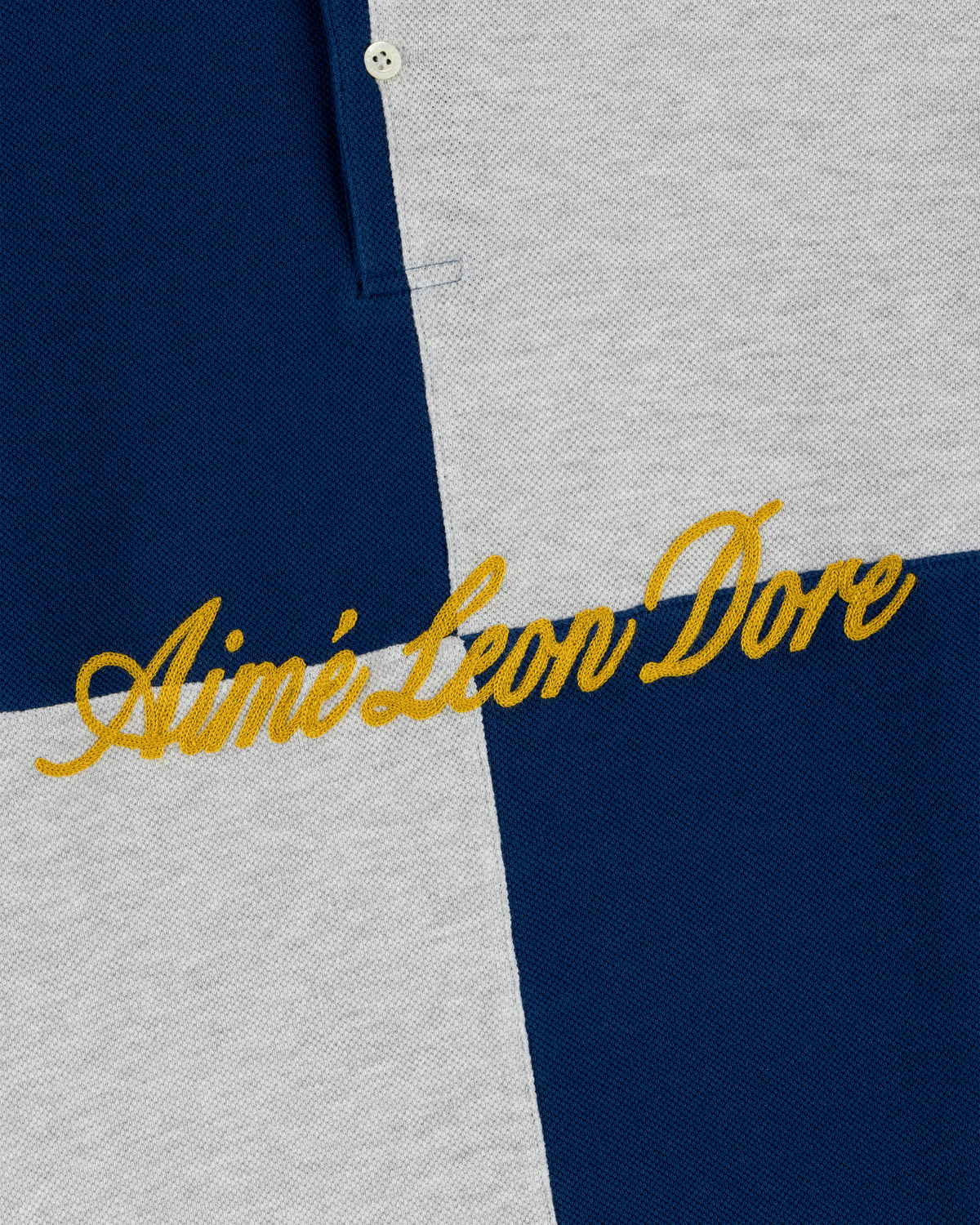 Close-up of the Color Blocked Pique Polo's blue and gray checkered fabric with yellow Aimé Leon Dore embroidery.