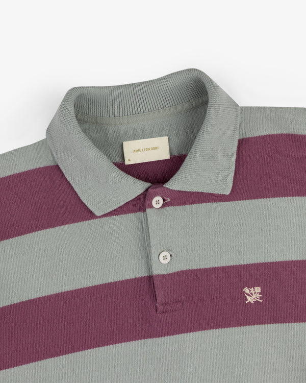 The Striped Crest Rugby is a gray and purple collared polo with two buttons and a small crest logo on the chest.
