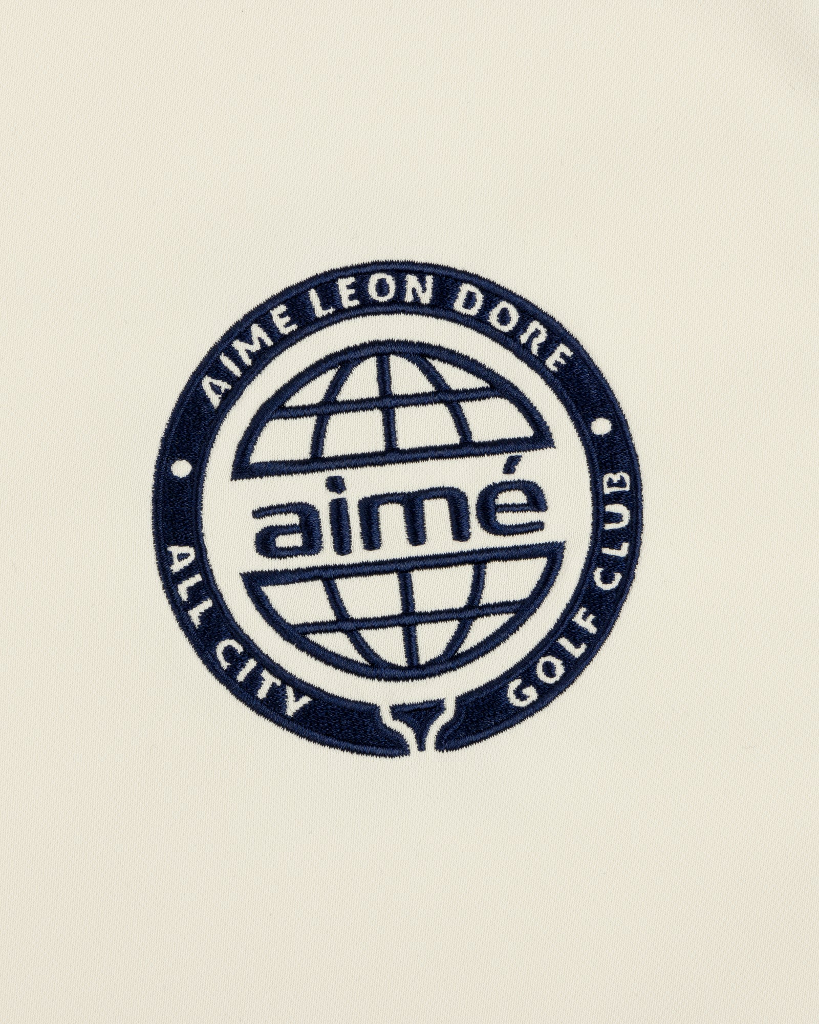 Navy blue ALD Golf Tour Polo with All City Golf Club globe logo and "aimé" on a cream background.