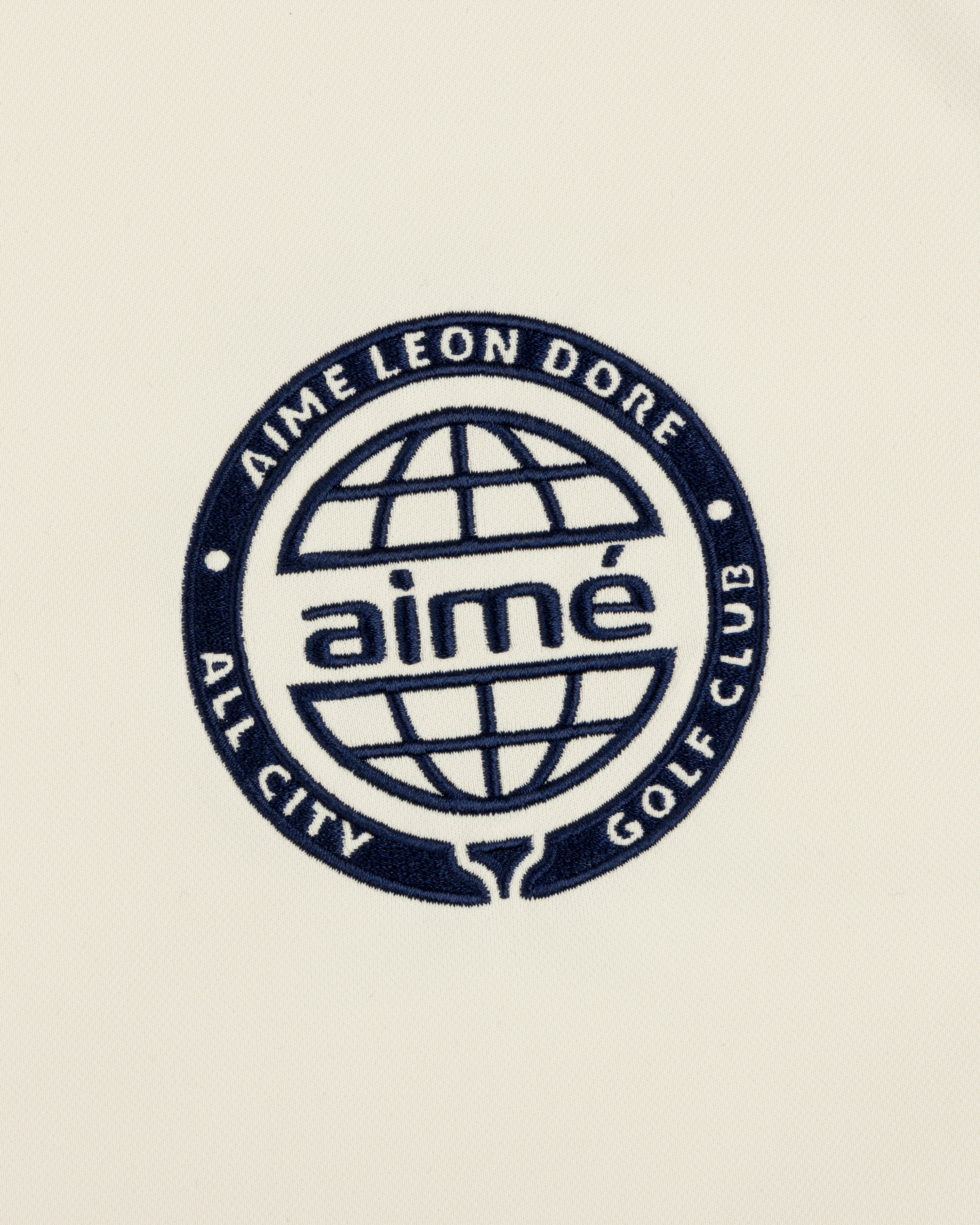 Navy blue ALD Golf Tour Polo with All City Golf Club globe logo and "aimé" on a cream background.