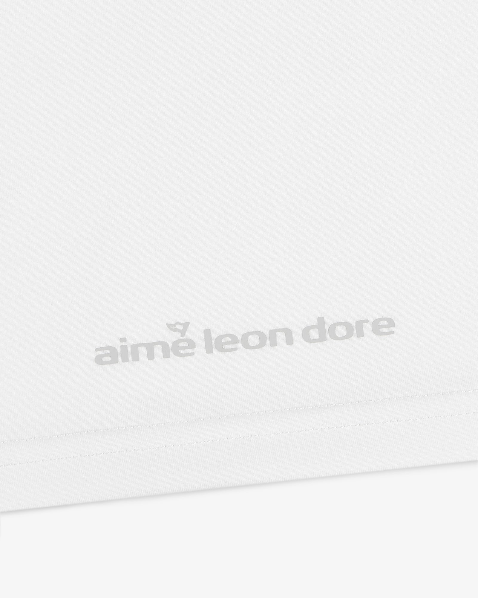 Close-up of a white ALD Golf Performance Tee with the aime leon dore logo in light gray near the bottom hem.