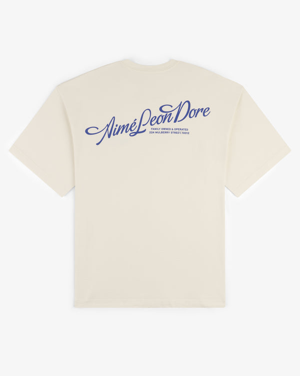 Beige Pappoús Logo Tee with Aimé Leon Dore logo and blue script text printed on the back near the shoulders.