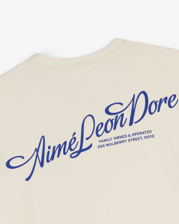 Pappoús Logo Tee: Beige shirt with large blue Aimé Leon Dore script and 224 Mulberry Street, 10012 on back.