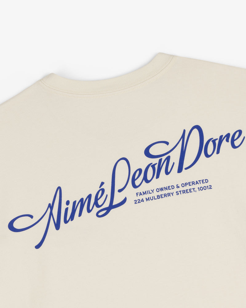 Pappoús Logo Tee: Beige shirt with large blue Aimé Leon Dore script and 224 Mulberry Street, 10012 on back.